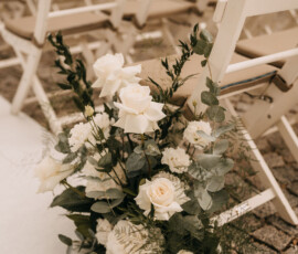 Romantischer Blumenschmuck für die freie Trauung: weiße Rosen, Eukalyptus und zarte Blüten sorgen für eine stilvolle Hochzeitsatmosphäre. Weiße Blumendekoration mit Rosen und Eukalyptus an Stühlen einer Hochzeitszeremonie im Freien.