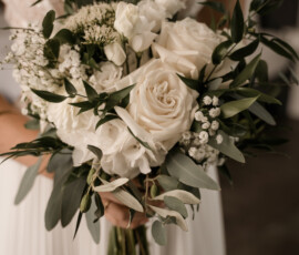 Zeitlos schön: Ein eleganter Brautstrauß mit weißen Rosen, Schleierkraut und Eukalyptus — liebevoll gestaltet von der Gärtnerei Reinwald. Braut hält einen weißen Brautstrauß mit Rosen, Schleierkraut und Eukalyptus vor ihrem Hochzeitskleid.