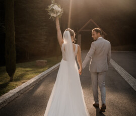 Ein Moment voller Glück: Das Brautpaar genießt den Tag Hand in Hand, begleitet von einem eleganten weißen Brautstrauß der Gärtnerei Reinwald. Brautpaar geht Hand in Hand im Abendlicht, die Braut hält ihren weißen Brautstrauß freudig in die Höhe.